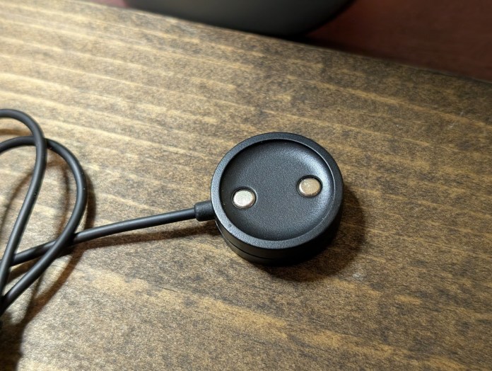 Close-up of a magnetic charging puck for a smartwatch, featuring two metal charging contacts and a connected cable, resting on a wooden surface.