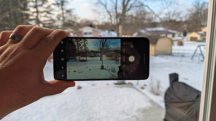 A person holding a smartphone to capture a photo of a snowy backyard scene through a window.