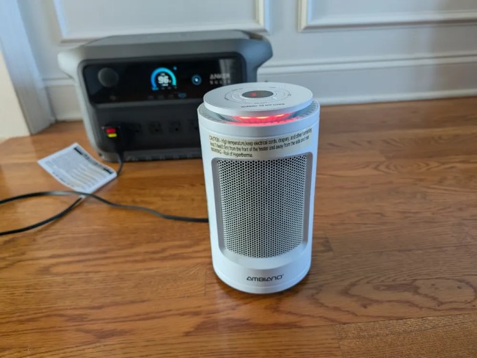 A compact white space heater with a digital control panel, featuring a red indicator light, positioned on a wooden floor near a power station and an instruction manual.